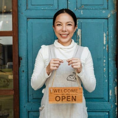 Cheerful ethnic female cafeteria owner in apron demonstrating cardboard signboard while standing near blue shabby door and windows after starting own business and looking at camera