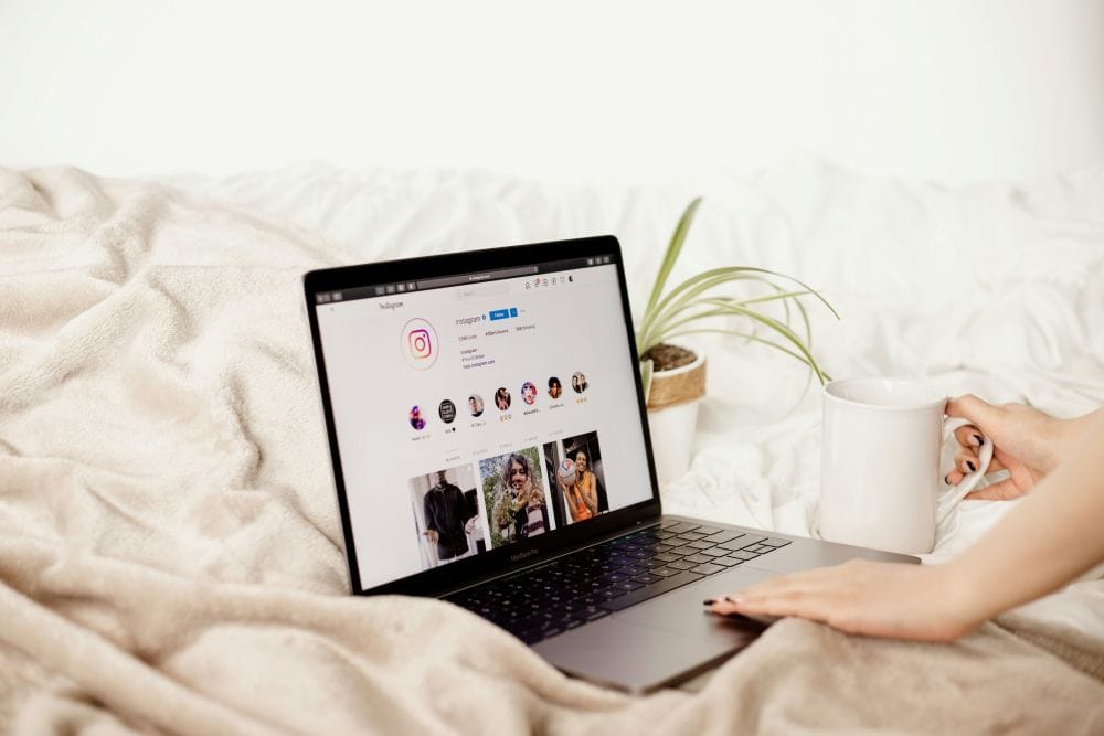 laptop on a bed with person's hands typing and holding coffee cup